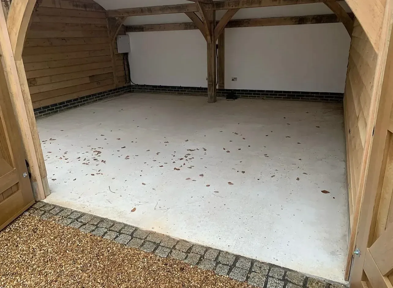 Empty oak-frame garage bay with bare concrete slab, leaves blown in, brick skirting and exposed timber framing before flooring